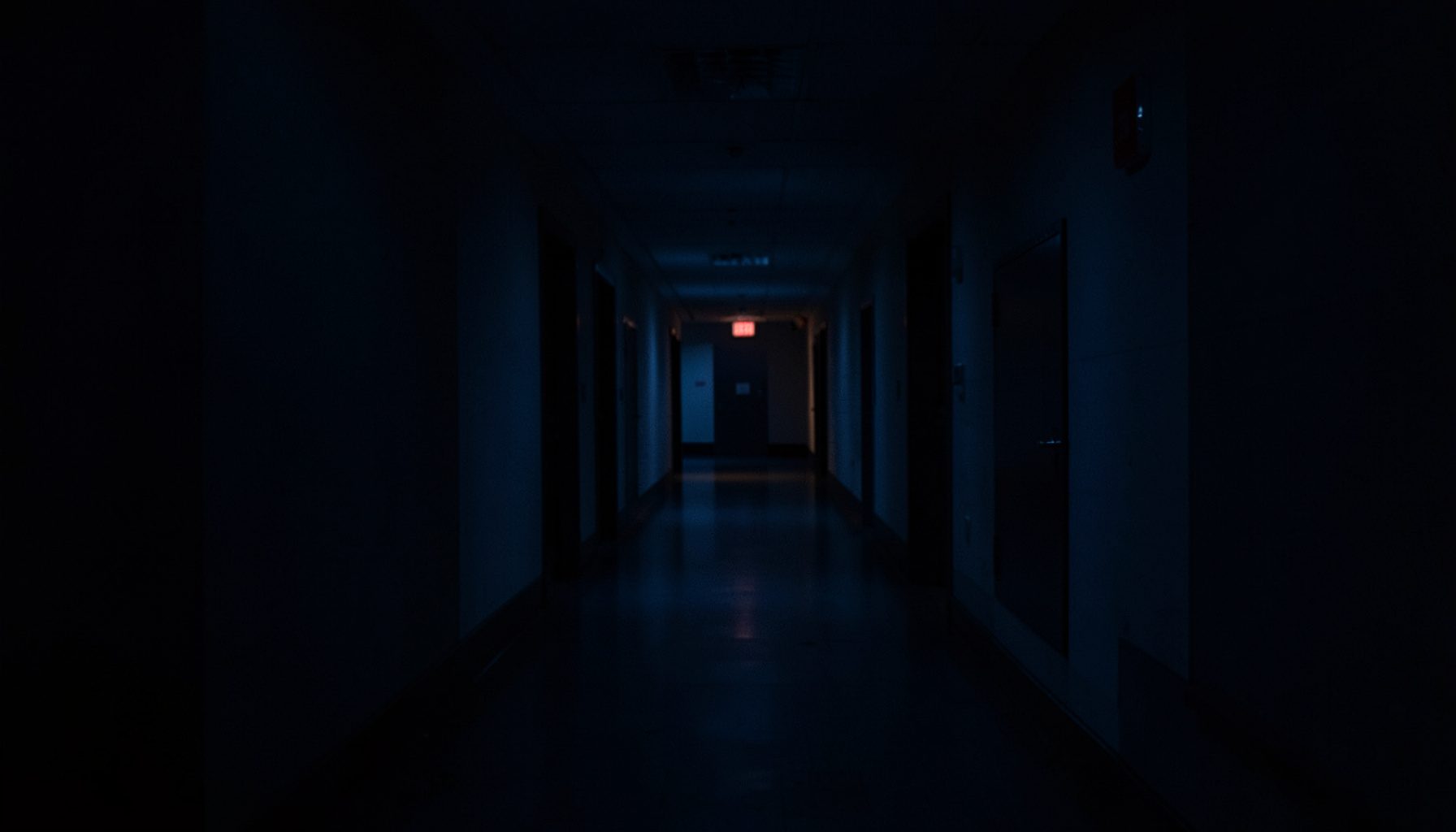 Dark low-key photograph of a quiet university corridor at dusk, deep navy shadows and a faint warm light at the far end