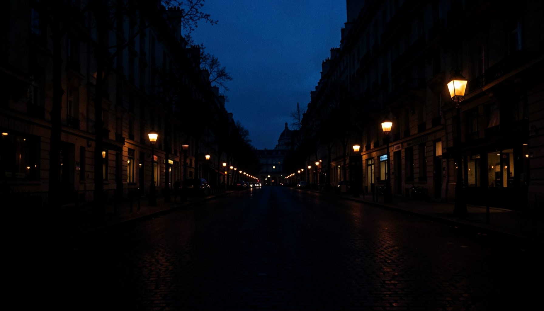 Low-key evening photograph of a wide Parisian boulevard in deep navy shadow, warm amber street lamps glowing faintly through the haze